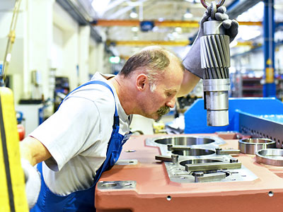Ein Mann in Arbeitskleidung untersucht ein Metallteil in einer Fabrik.